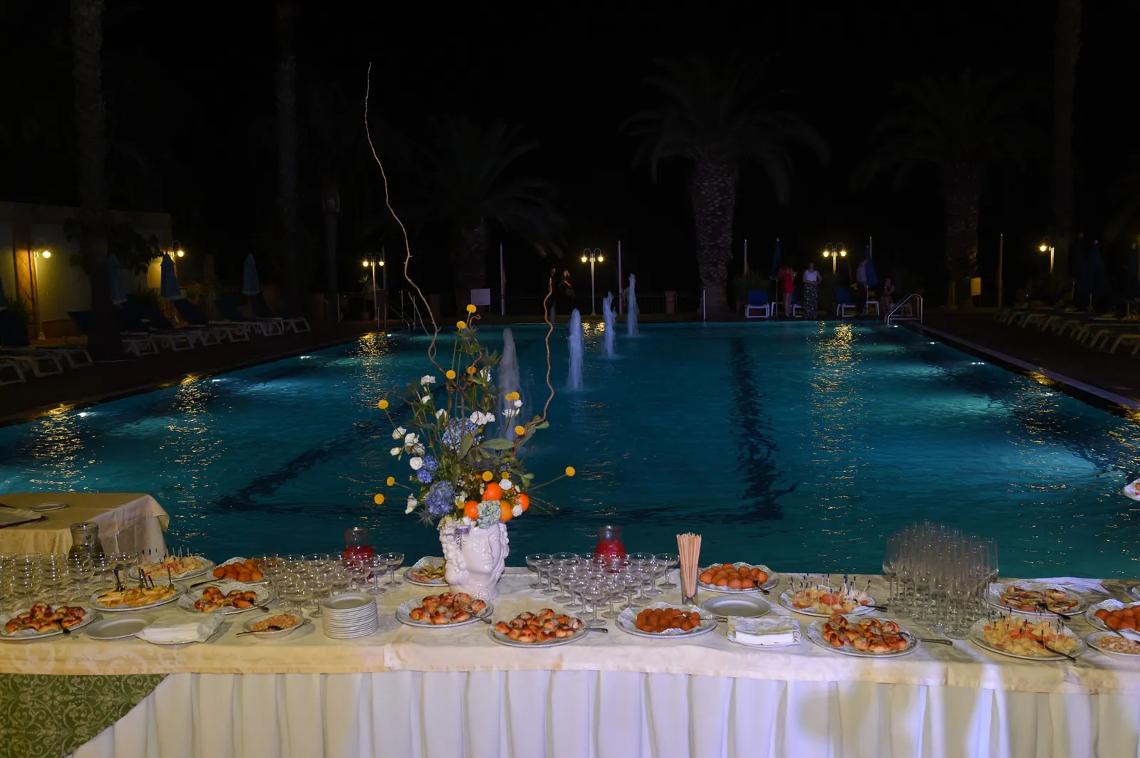 La piscina illuminata di notte con i getti d'acqua e, in primo piano, il bordo della tavola imbandita per il banchetto