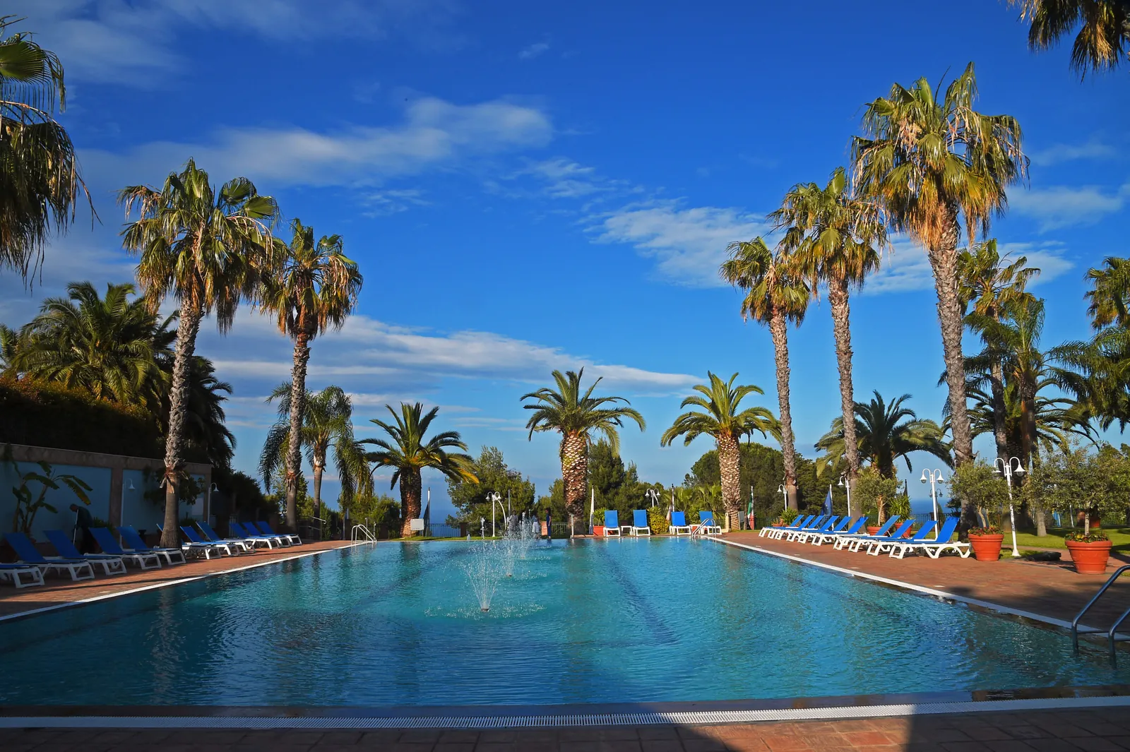 La piscina scoperta con i getti d'acqua, i lettini azzurri e le palme; il bordo dell'acqua sta sulla linea dell'orizzonte